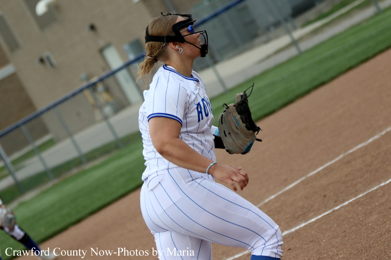 Side view of a female softball pitcher in a white pinstripe uniform on the mound, mid-throw with a gloved hand and protective mask nearby.