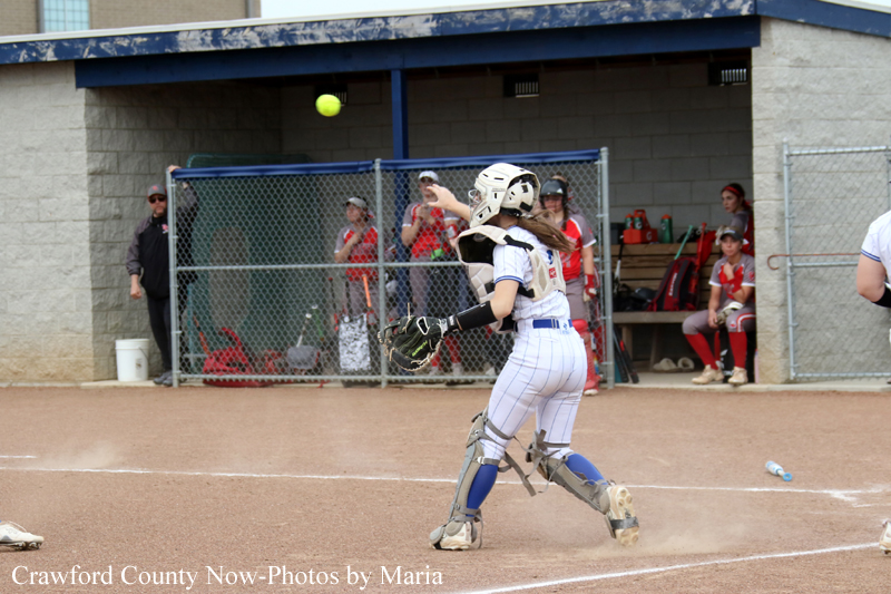 Female softball catcher in a white uniform and full protective gear reaches to catch a ball in midair near home plate, dugout in the background.