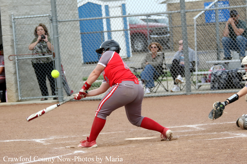 Softball batter in red and gray swings at a pitched ball on a dirt field, with fence and spectators behind.