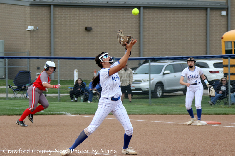 Softball pitcher in white and blue winds up a throw on the infield as a red-uniform runner advances and a teammate waits near first base.