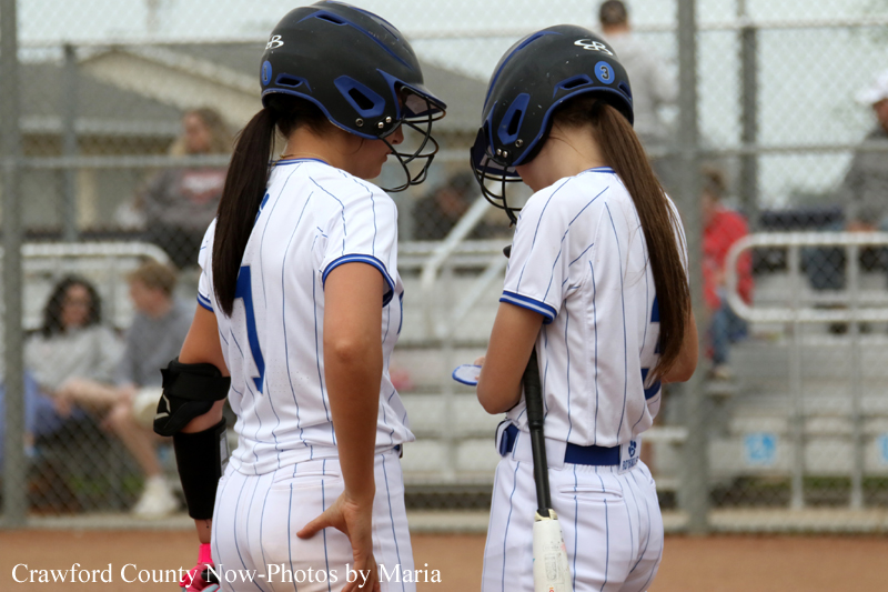Two young softball players in white pinstriped uniforms with helmets stand facing each other with bats at a field.