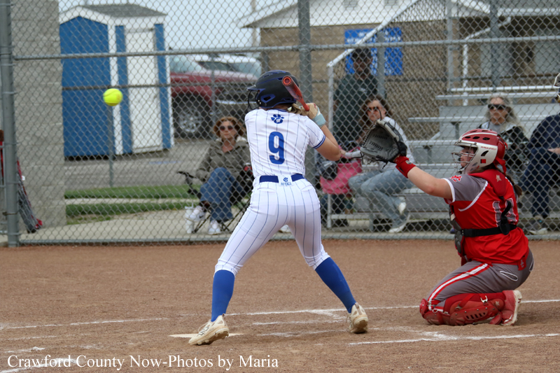 Female softball batter in a white pinstripe uniform with number 9 swings at a pitch as a red-gear catcher crouches behind home plate; ball in midair near the plate.