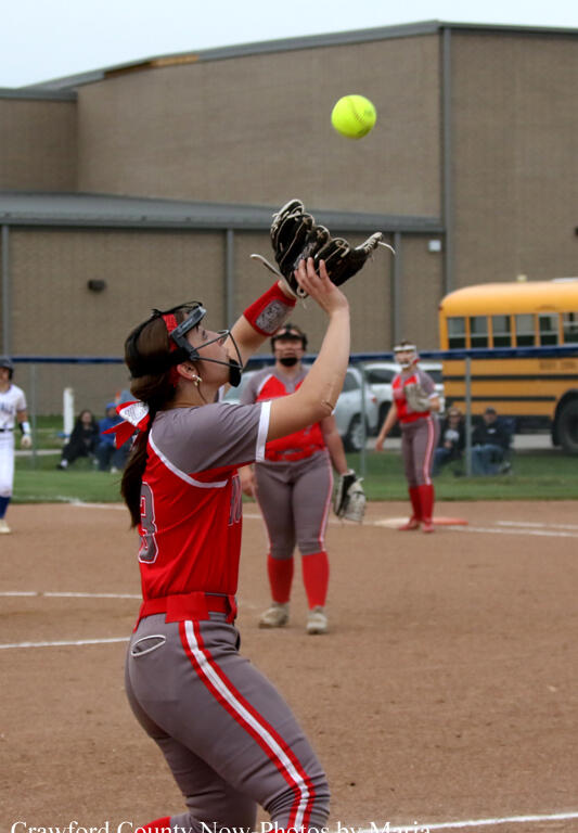 Softball player in red and gray uniform reaching up with a glove to catch a yellow ball on the infield; teammates and a school bus in the background.