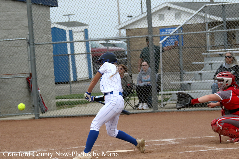 Baseball batter in white pinstripes swinging at a pitch as catcher in red crouches behind home plate behind a chain-link fence.