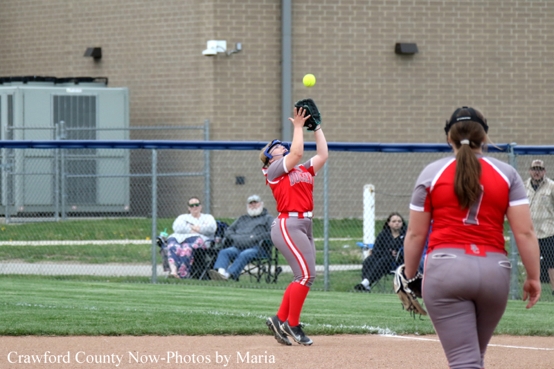 Softball player in red uniform leaps to catch a high ball with her glove while another fielder stands nearby on the infield; spectators sit behind a chain-link fence beyond the outfield.