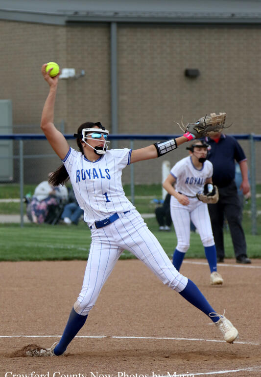 Female softball pitcher in a white pinstripe Royals uniform throws from the mound, with a teammate in the background.