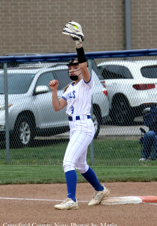 Youth softball player in a white pinstriped uniform stands on a base with a glove raised overhead, signaling during a play.