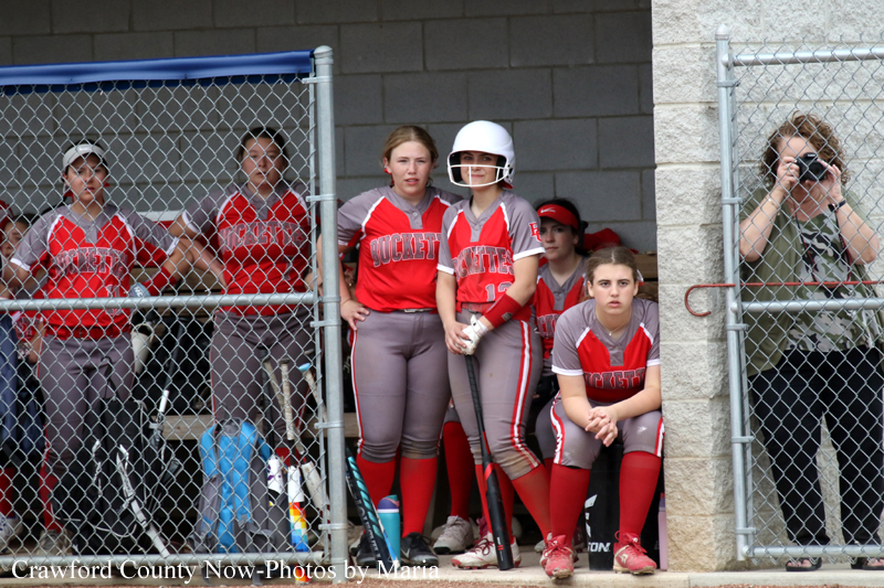 Softball team in red and gray uniforms sits and stands in a dugout behind a chain-link fence, some holding bats and wearing helmets.