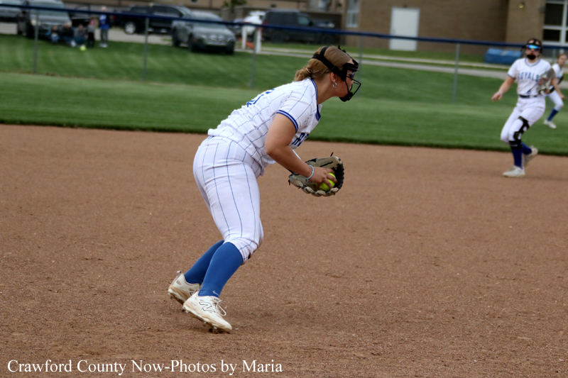 Softball player in white pinstripe uniform and blue socks fields a ground ball on the infield, wearing a helmet and glove; teammate in the background.