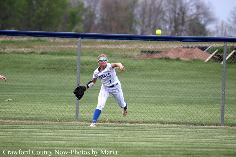 Female softball player in white and blue uniform reaches for a ball near the outfield fence during a game.