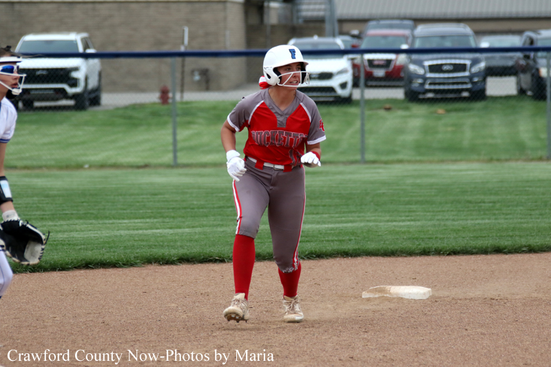 Young softball player in red and gray uniform runs between bases on the dirt infield, helmet on, with a white base nearby and a fielder partially visible on the left.