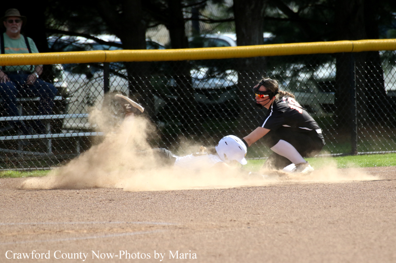 Softball scene: a baserunner in a white helmet slides into a base as a fielder in a black jersey reaches to tag, kicking up a large cloud of dust. Behind them is a chain-link fence and spectators.