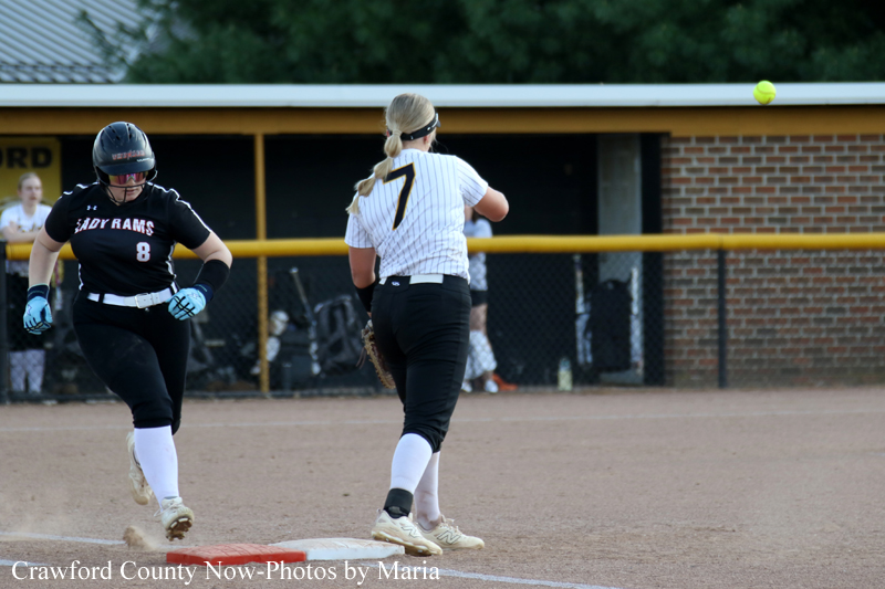 Two female softball players near a base: a white-jersey runner (7) and a black-jersey fielder (8) with a ball in the air nearby in the infield roadbed area.