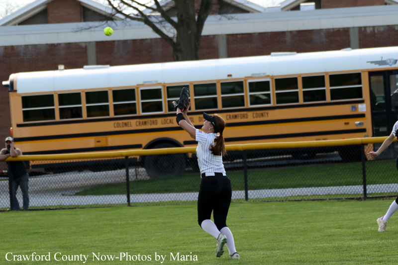 Female softball player in white uniform leaps to catch a ball with a glove on a grassy field; yellow school bus and fence in the background.