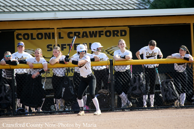 Softball batter in a white uniform swings at a pitch while teammates watch from the dugout behind the fence.