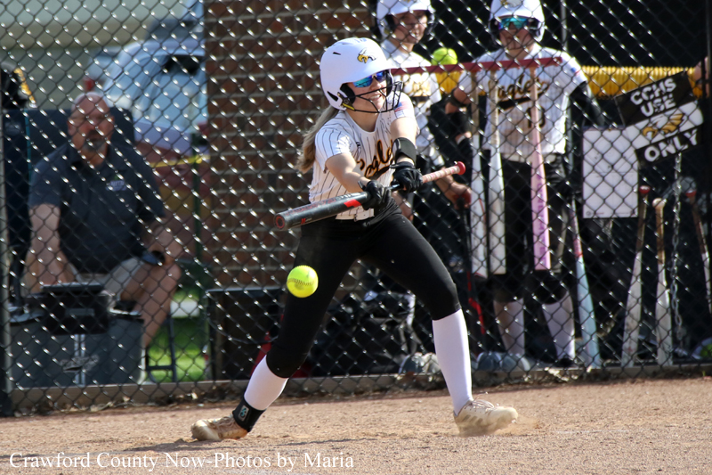 Female softball batter swings at a pitched ball near home plate, wearing white helmet and black pants, behind a chain-link fence.