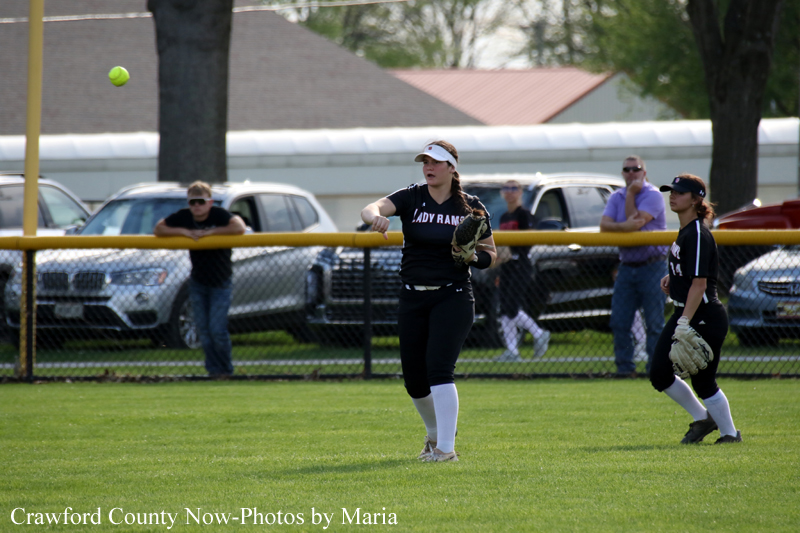 Softball game action: pitcher in a black uniform throws a pitch as a fielder watches, with spectators behind a chain-link fence.