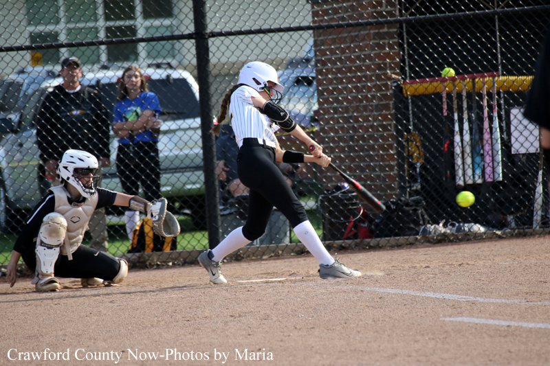Female softball batter swings at a pitch as the catcher crouches behind home plate on a dirt field with a chain-link fence in the background.