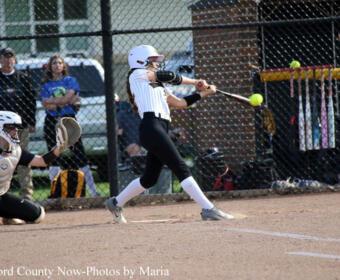 Female softball batter swings at a pitch near home plate as catcher crouches behind her on the infield dirt.