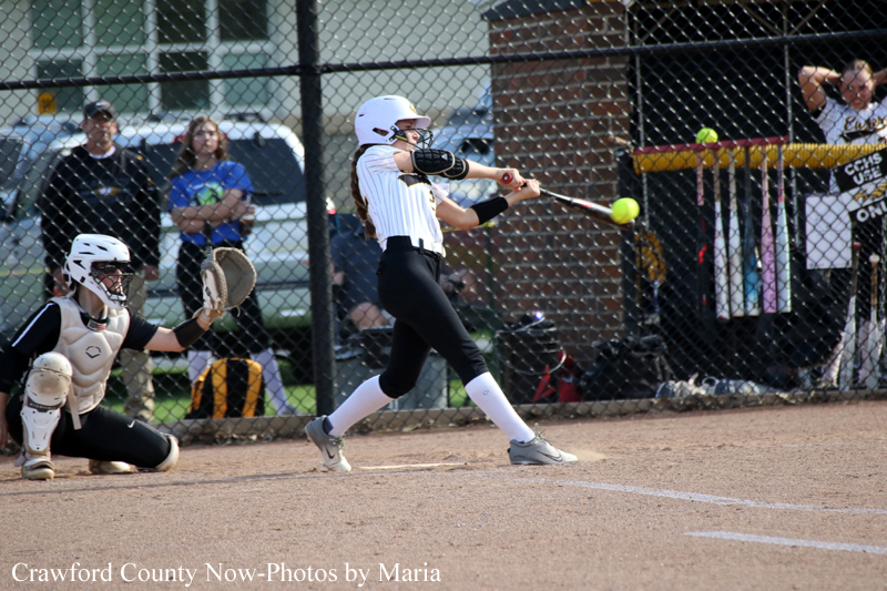 Female softball batter swings at a pitch near home plate as catcher crouches behind her on the infield dirt.