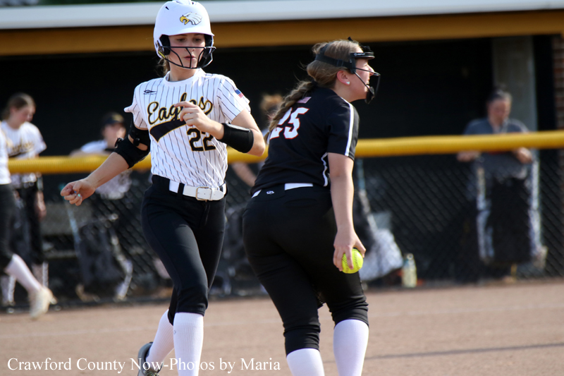Female softball players in action on a dirt field; runner in white pinstripe uniform (#22) and catcher in black (#25) near the bases as a yellow ball is visible nearby.