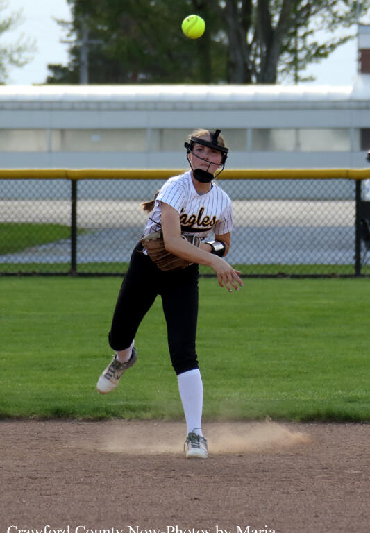 Female softball pitcher in a white Eagles jersey, mid-pitch on a dirt mound with dust rising, wearing helmet and glove on a sunny field.