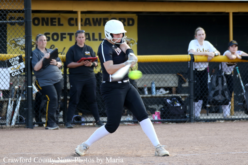Female softball batter in a white helmet and black uniform swings at a pitch on a dirt field, with a chain-link fence and spectators in the background.