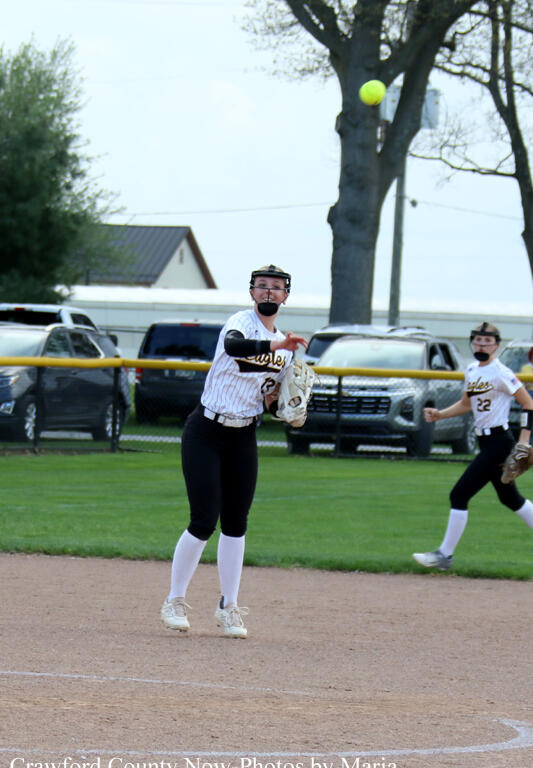 Female softball pitcher in a white pinstripe uniform delivers a pitch on a dirt mound, glove ready, with a teammate nearby on the field behind her.