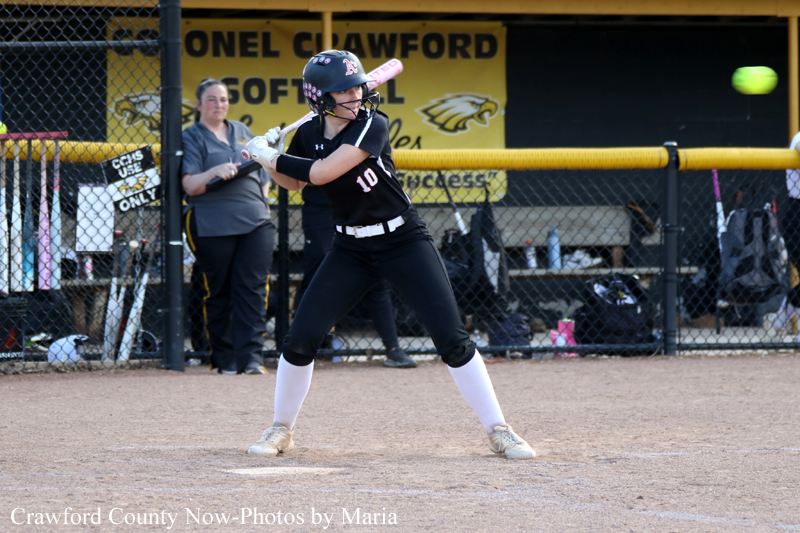 Softball batter in black uniform #10 swings at a pitch on a dirt field, helmet on, with a chain-link fence and a yellow banner in the background.