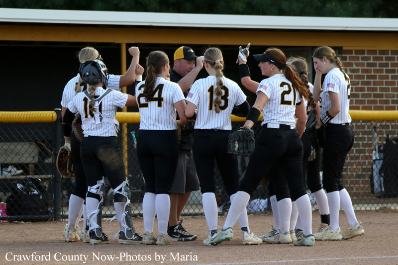 Softball players in a circle huddle on the infield, wearing numbered uniforms, talking to a coach during the game.