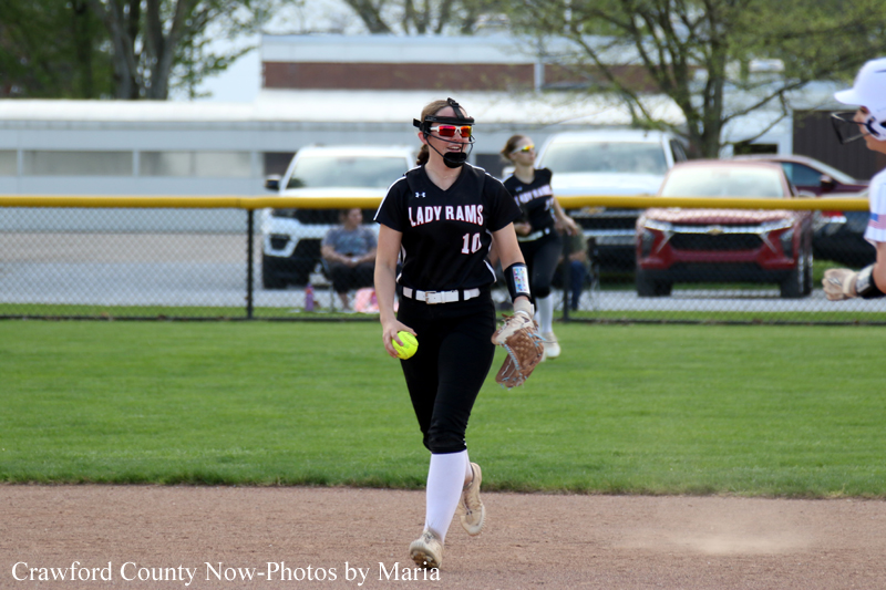 Softball player in a black Lady Rams uniform (number 10) runs on the infield with a glove, holding a bright yellow ball in her other hand.