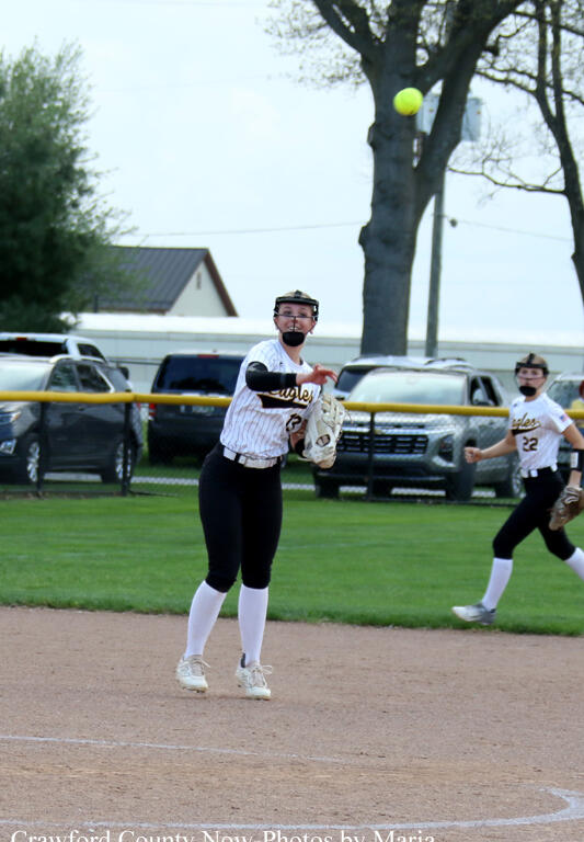 Female softball pitcher in a white pinstripe uniform throws a pitch on the infield, neon ball in midair behind her glove
