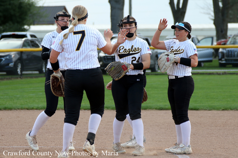 Four female softball players in white pinstripe uniforms celebrate with a high-five on the infield, gloves on, with 'Eagles' on their jerseys and numbers visible (one player with 7).