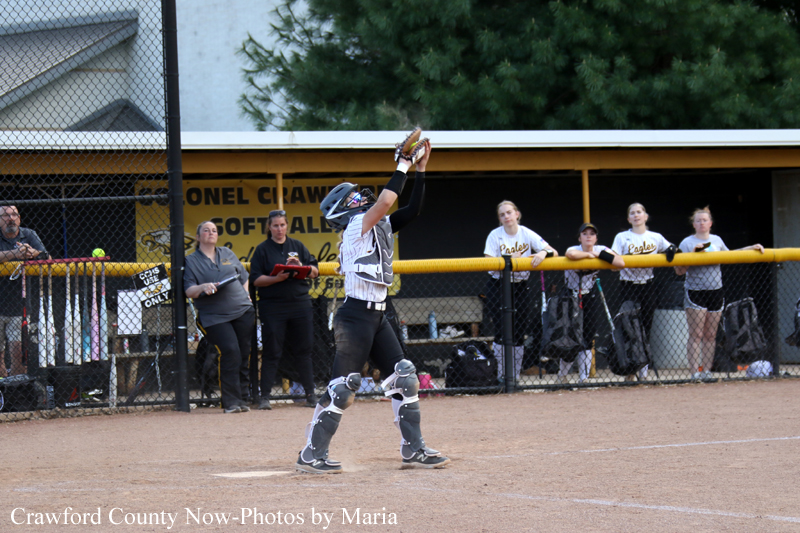 Softball catcher in protective gear reaches overhead to catch a high throw at home plate, with teammates watching behind the fence.