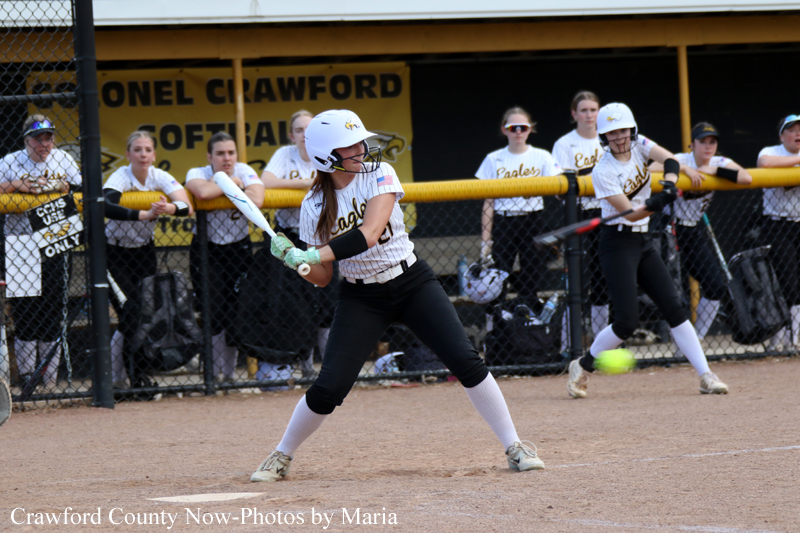 A softball batter in a white helmet swings at a pitch while teammates watch from the dugout behind a chain-link fence.