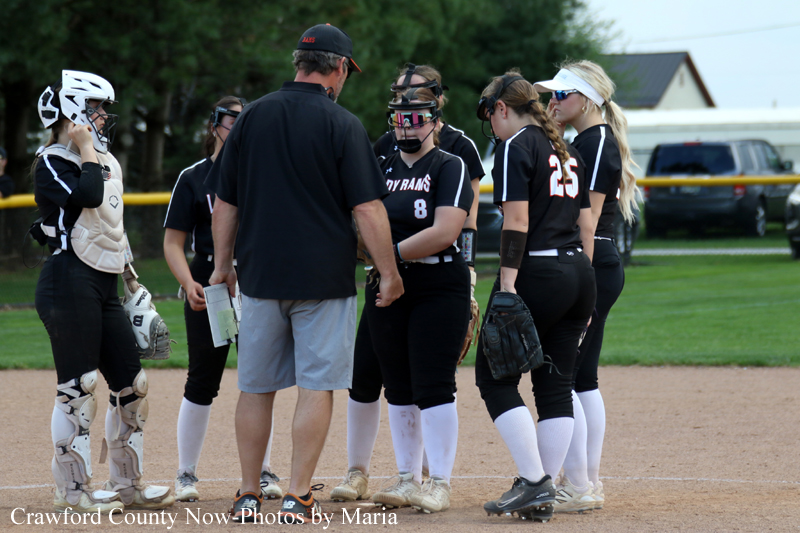 A group of female softball players in black uniforms listening to their coach on the infield, with a catcher in gear on the left.