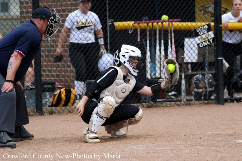 Softball catcher in white helmet and protective gear diving to block a ball near home plate with an umpire watching in the background.