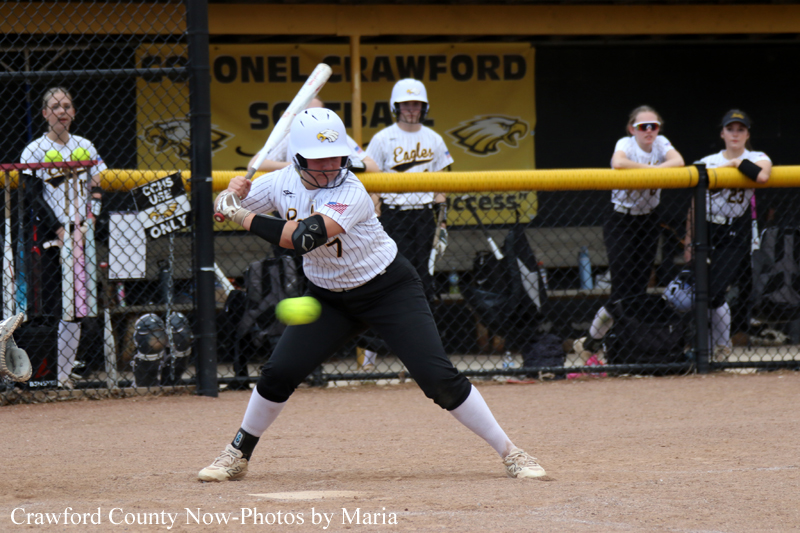 Female softball batter in a white pinstripe uniform swings at a pitch, helmet on, near home plate on a dirt field with players watching from the dugout behind a chain-link fence.