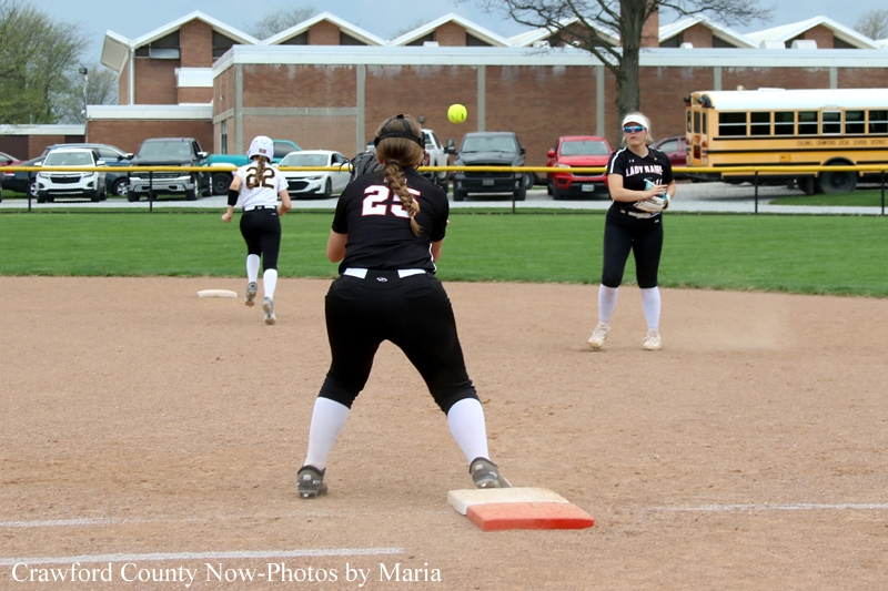 Softball infield action: a fielder in black (number 24) on first base as the ball is pitched, with a runner approaching from the left and a keeper on the field; buses and cars in the background.