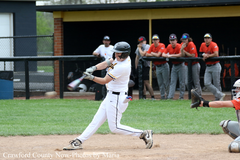 Baseball batter in a white uniform swings at a pitch as the catcher and a row of players watch from the dugout in the background.
