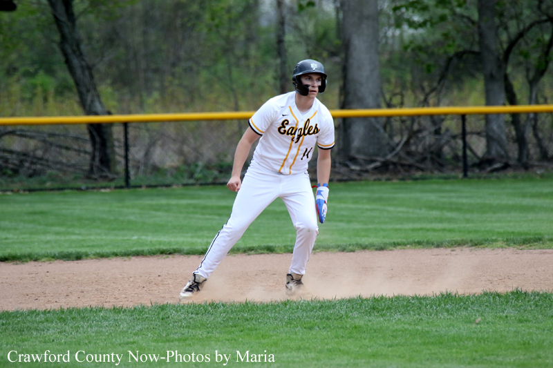 Baseball player in a white Eagles uniform, helmet on, standing on the infield dirt during a game with green grass and trees in the background.
