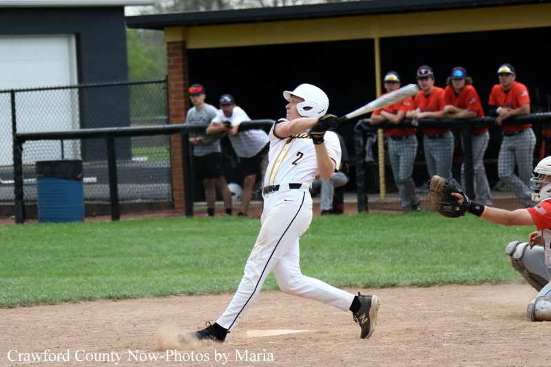 Baseball batter in a white uniform swings at a pitch while the catcher in orange reaches with his glove behind the plate, with teammates watching from the dugout in the background.