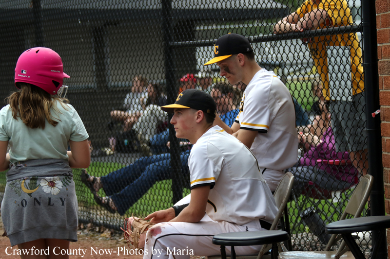 Baseball players in white uniforms sit on benches behind a chain-link fence while a girl wearing a pink batting helmet stands nearby.