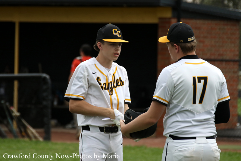 Two baseball players in white and gold Eagles uniforms shake hands on a field near a dugout, one facing the camera wearing a CC cap.