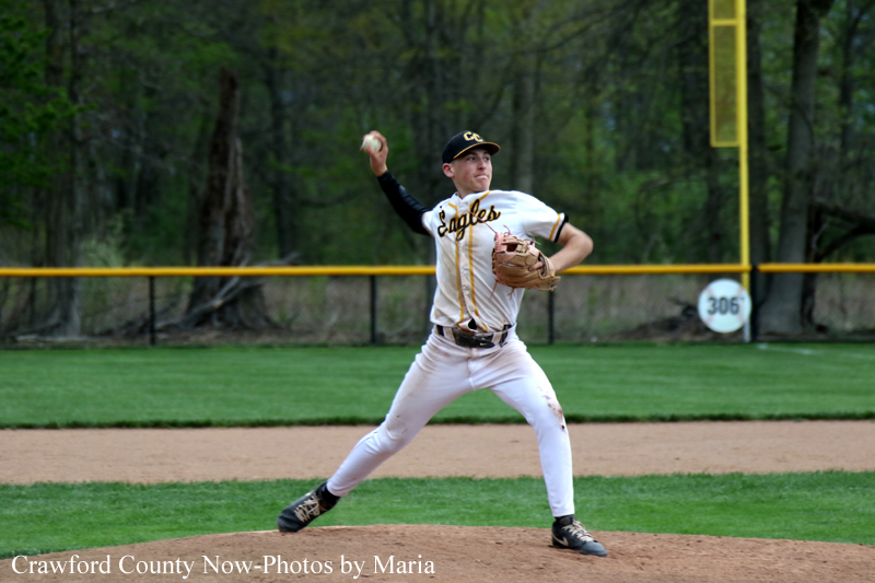 Baseball pitcher in white-and-gold uniform winding up on the mound, Eagles on jersey, with outfield fence and trees behind him.