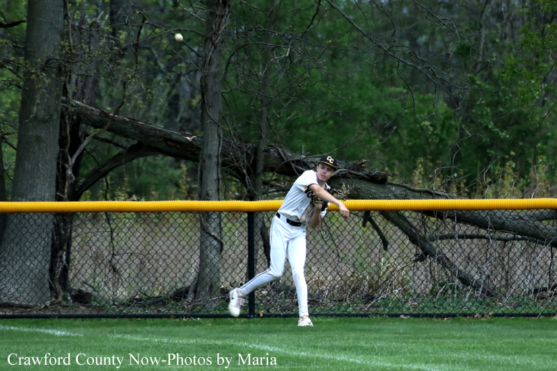 Baseball player in a white uniform and brown cap fields a ball near the outfield fence on a grassy field.