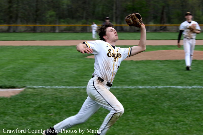 Action shot of a baseball pitcher in a white and yellow Eagles uniform mid-throw on a grassy field, with teammates in the background.
