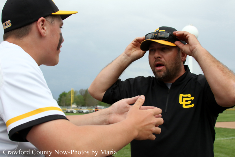 Two men on a baseball field talk while one adjusts his cap; one wears a black shirt with a yellow logo.