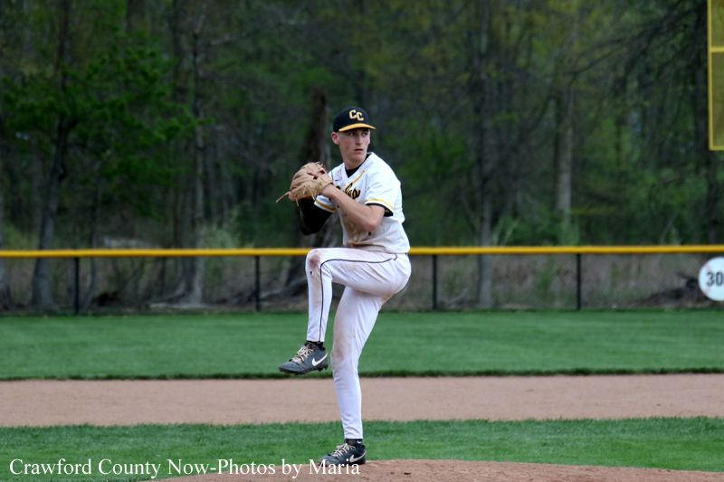 A baseball pitcher in a white uniform winds up on the mound during a game, glove raised and ready to throw, with a green field and outfield fence in the background.