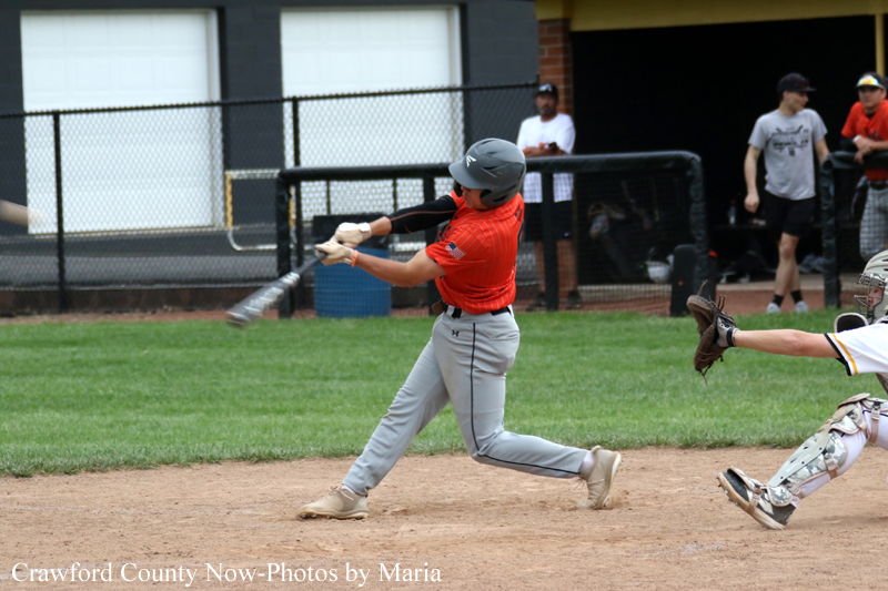 Baseball player in a red jersey swings a bat during a game, with a catcher reaching from the right in the dirt and grass field.